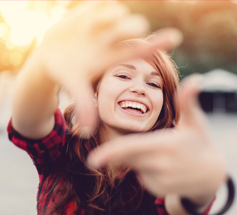 A young woman with long red hair and a black and red flanel shirt laughs directly into the camera, forming a square with both her forefingers and thumbs.
